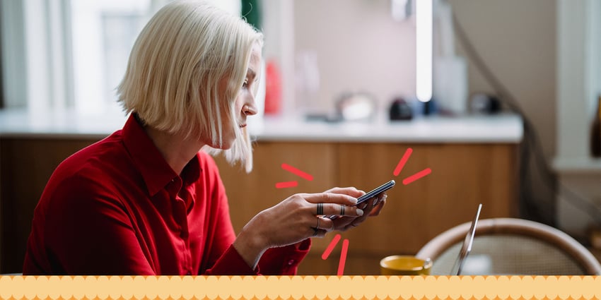 A woman with short blonde hair, wearing a red shirt, sits at a table using her phone. A laptop and coffee cup are on the table in front of her.