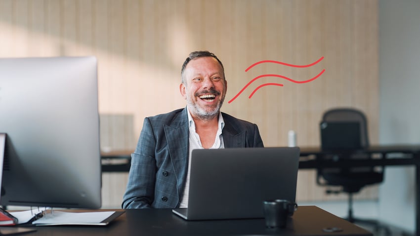 Joyful man with a beard, laughing at his desk in a modern office, with a laptop and a cup visible.