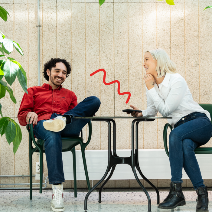 Two colleagues sitting and laughing at a small table in a bright, modern office with plants, one holding a phone.