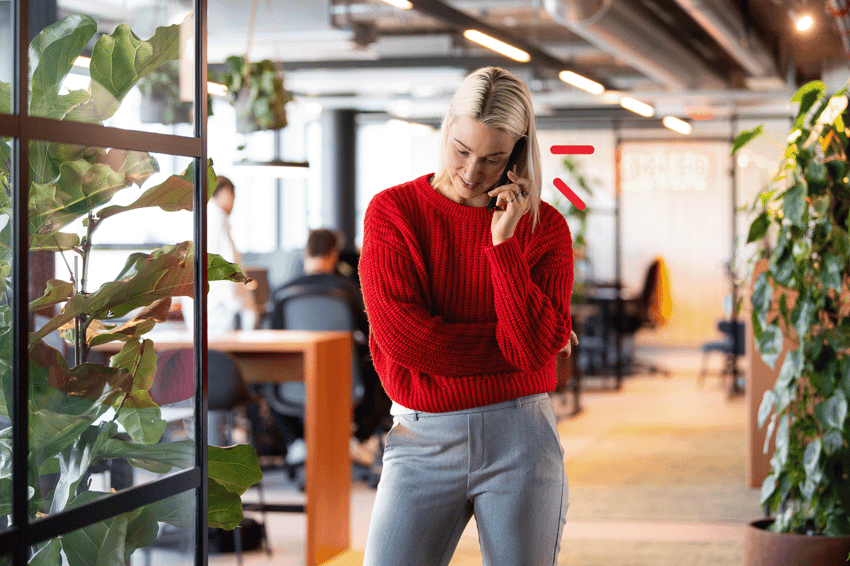 A woman in a red sweater smiling while talking on her phone, standing in a vibrant office space with plants and natural light.