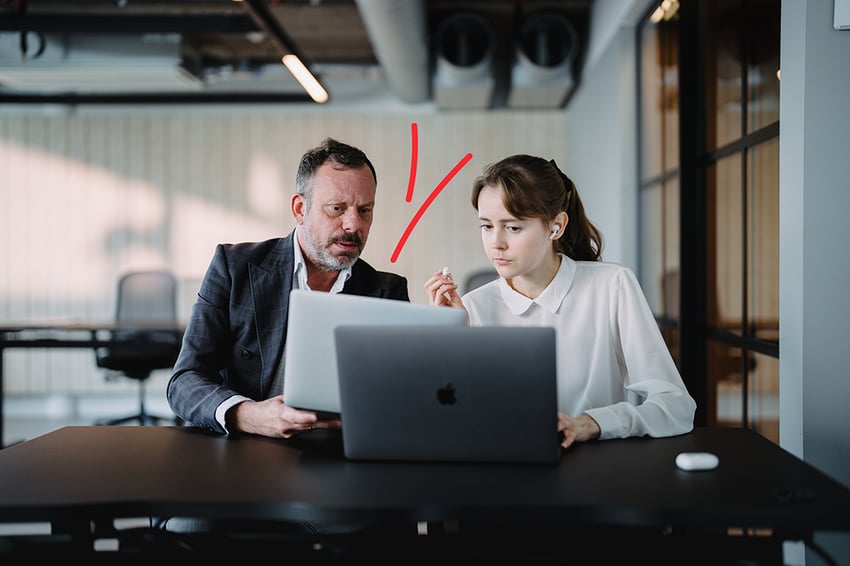 A focused young woman and an older man in a suit review content on a laptop in a modern office setting.